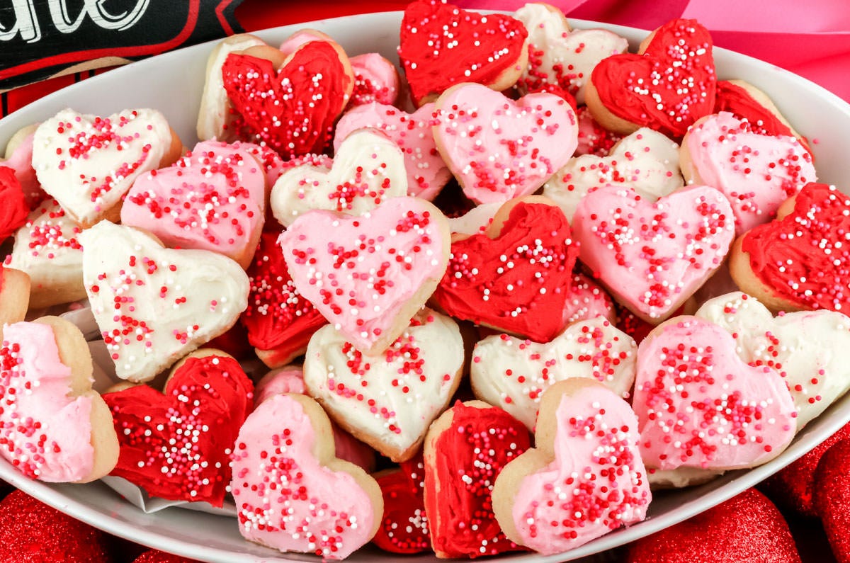 A plate of pink and red frosted sugar cookies, shaped like hearts and covered in red and white sprinkles.