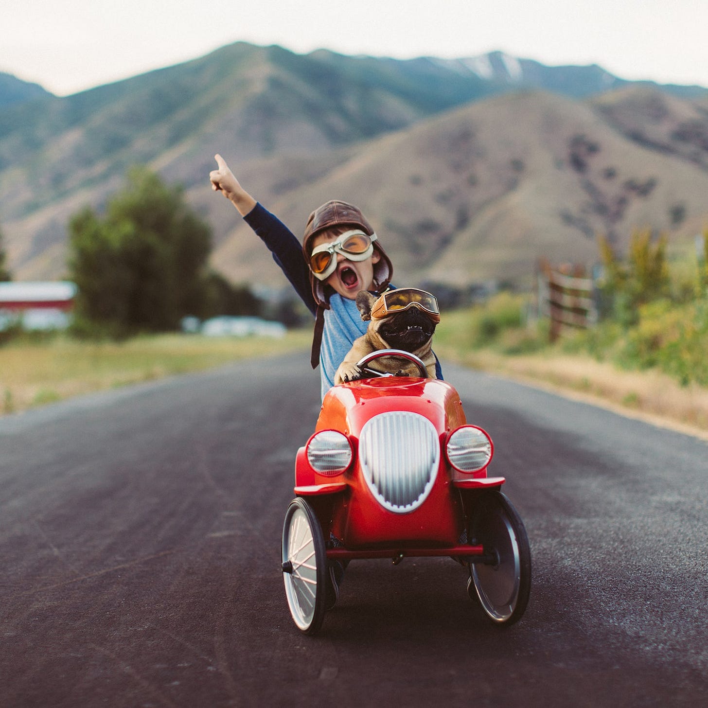 A young boy with flying goggles and flight cap races a red toy car with his pet and best friend French Bulldog along a small road in Utah. A young boy with flying goggles and flight cap races a red toy car with his pet and best friend French Bulldog along a small road in Utah.