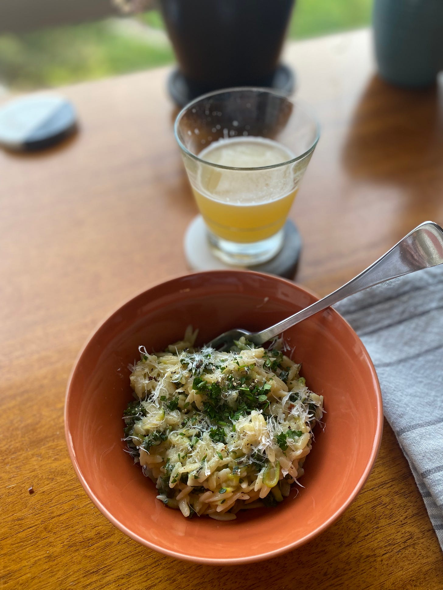 An orange bowl of the orzo described above, with pieces of kale and zucchini visible throughout the pasta. On top is grated parmesan and chopped parsley. A striped grey napkin is on the right side of the frame.