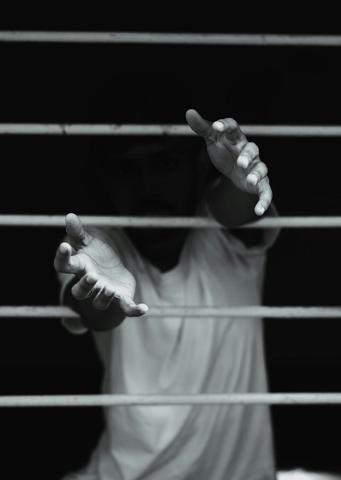 A figure in shadow reaches his hands through the bars of a prison cell, as if begging for help from those standing outside. A figure in shadow reaches his hands through the bars of a prison cell, as if begging for help from those standing outside.