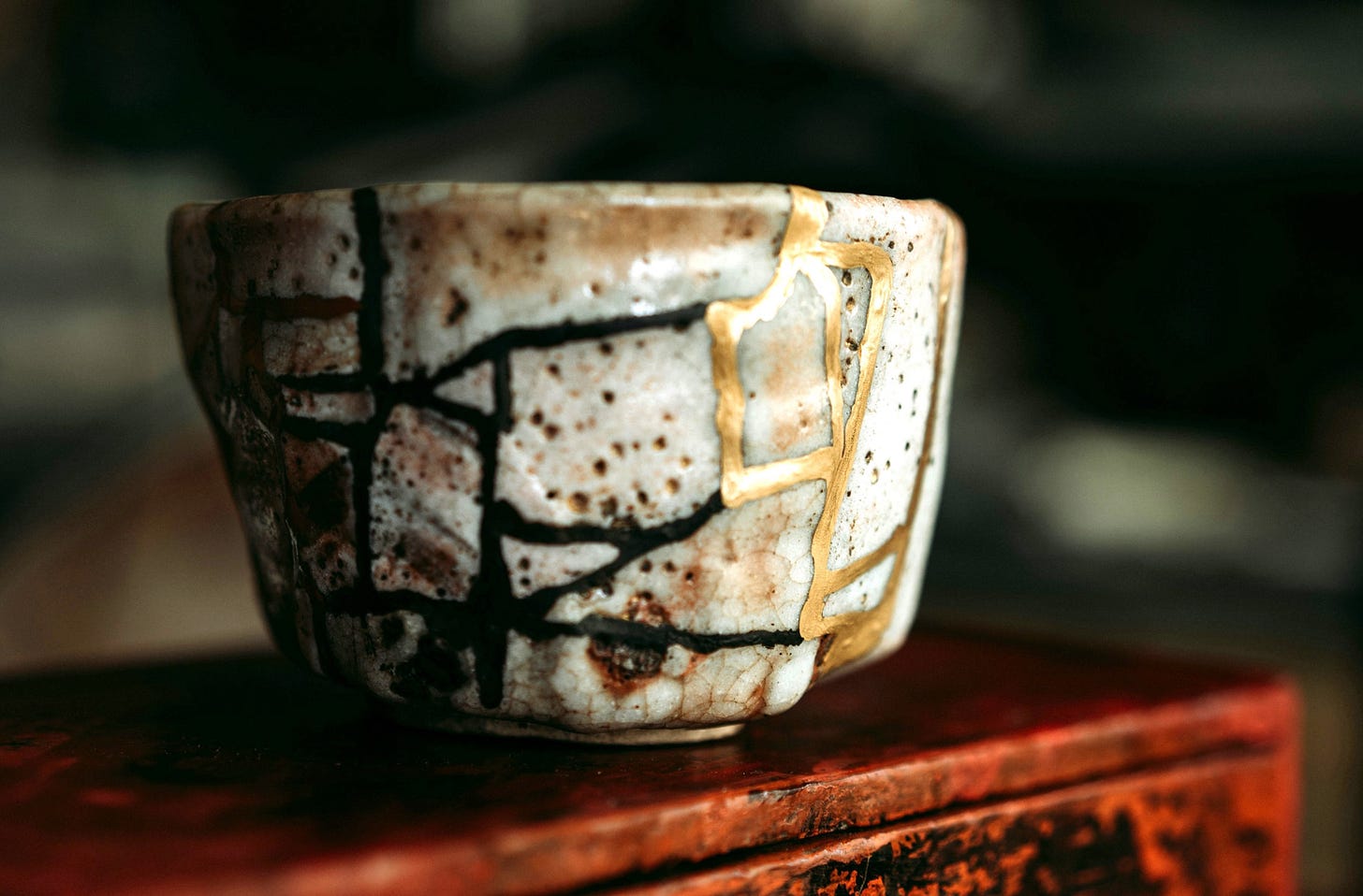 small white ceramic bowl with gold and black fracture lines. It is sitting on the edge of a red-brown wooden table.