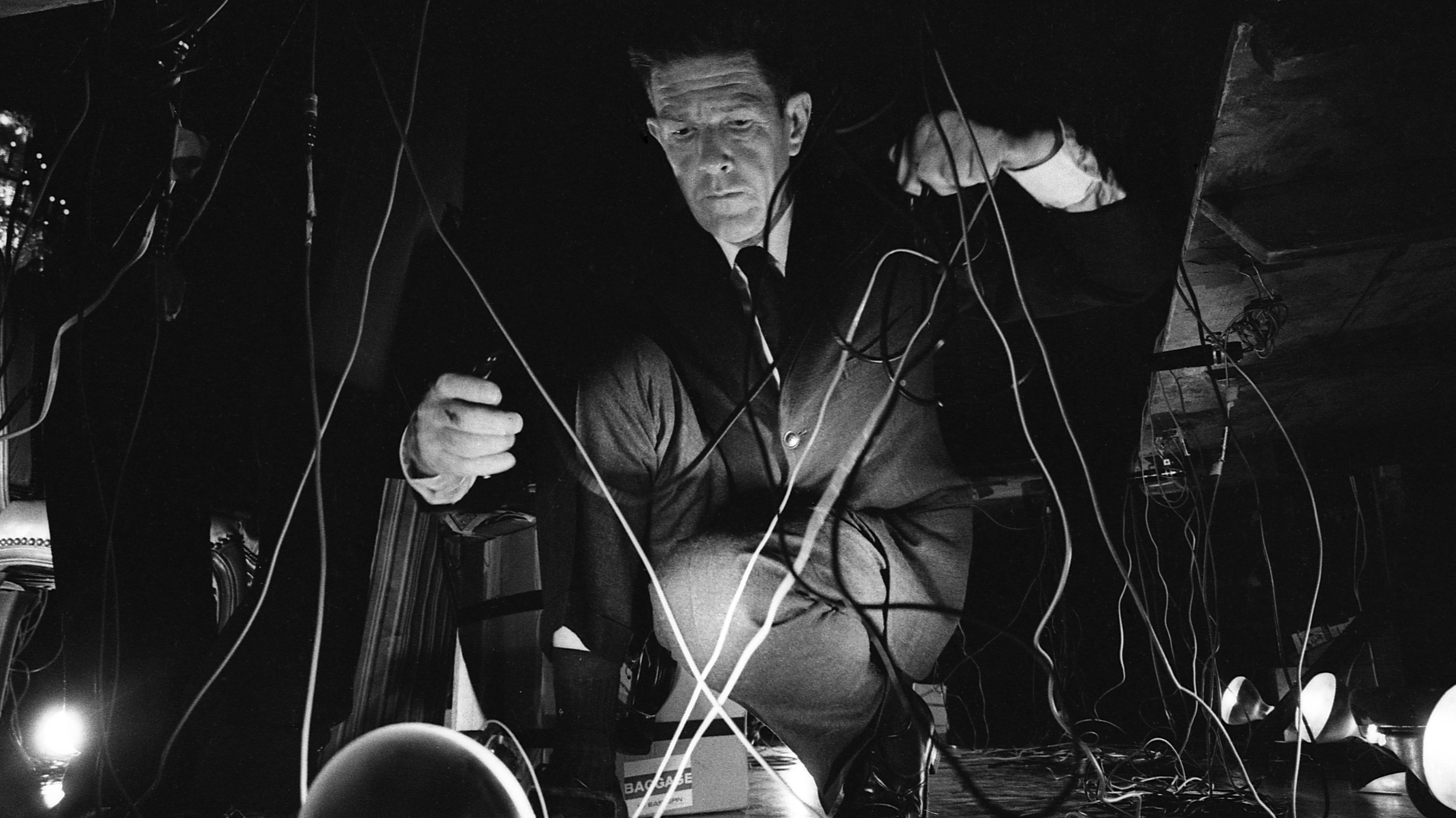 Black and white photo of composer John Cage crouched among a tangle of wires and stage lights, focused intently as he prepares an experimental performance.