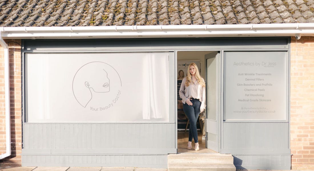 A woman standing in front of a grey building front that says 'Your Beauty Doctor'