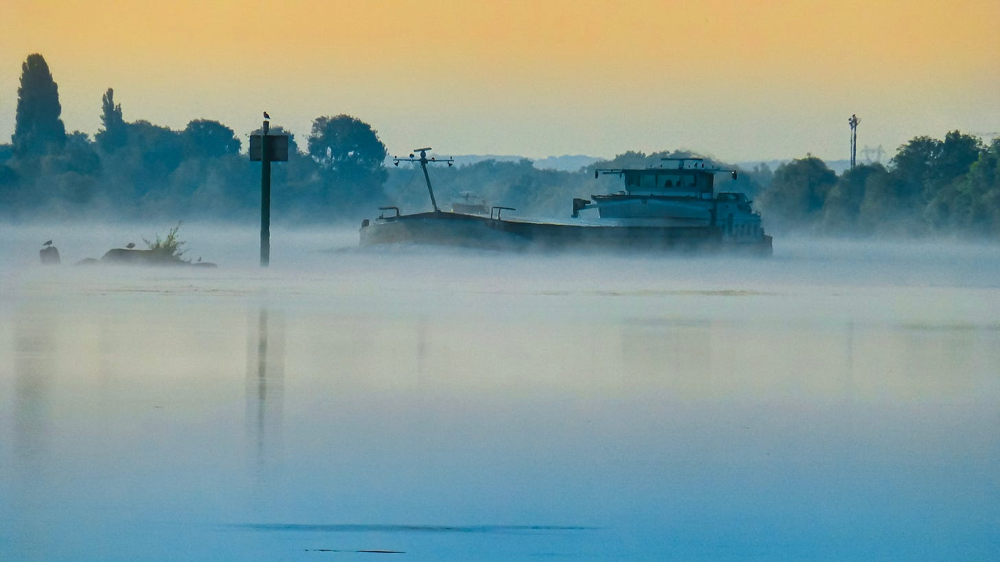 Large barge emerging through river mist at sunrise, trees and sky softly visible in the background.
