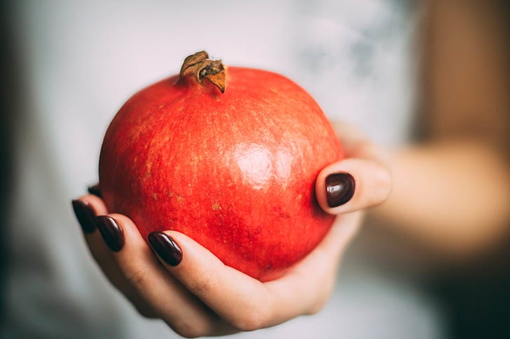 Picture shows a pomegranate in a pale-skinned hand with dark brown nail polish - the background is blurred, appearing to show the torso and arm of a person wearing white thrusting the fruit at the viewer Picture shows a pomegranate in a pale-skinned hand with dark brown nail polish - the background is blurred, appearing to show the torso and arm of a person wearing white thrusting the fruit at the viewer