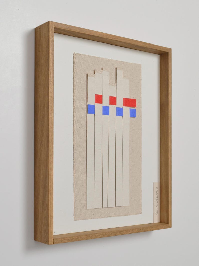 May include: Abstract art piece in a wooden frame. The artwork features vertical cream-colored strips with red and blue squares at the top, set against a beige canvas background. The frame is a light brown color.