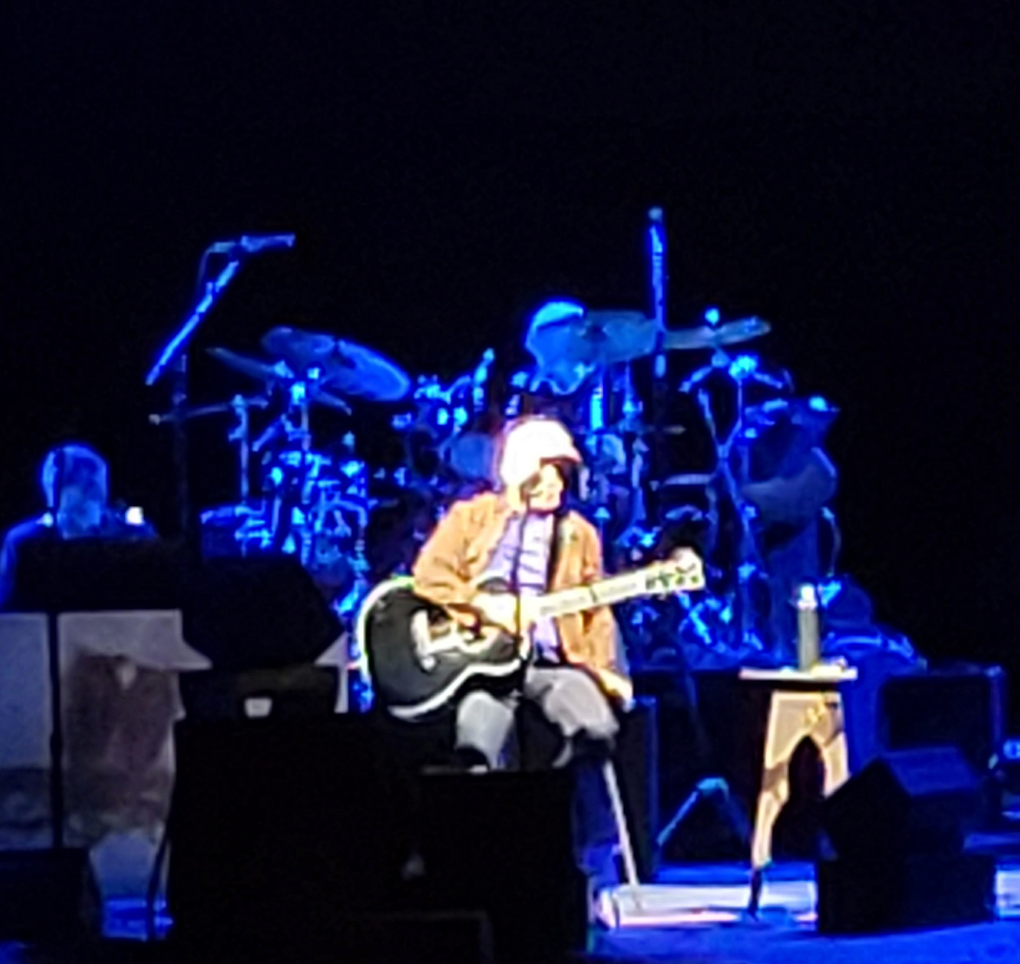 A blurry photograph of Paul Simon with guitar onstage and the rest of the band bathed in blue light