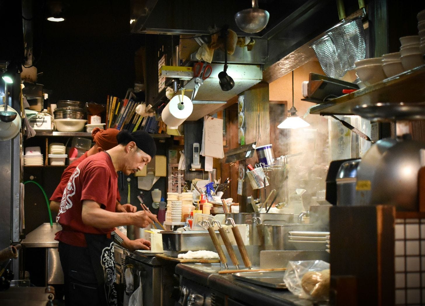 A cook working in a small Japanese ramen shop, steam rising under warm light, surrounded by stacked bowls and worn utensils—the kind of sensory moment that surfaces months later. A cook working in a small Japanese ramen shop, steam rising under warm light, surrounded by stacked bowls and worn utensils—the kind of sensory moment that surfaces months later.
