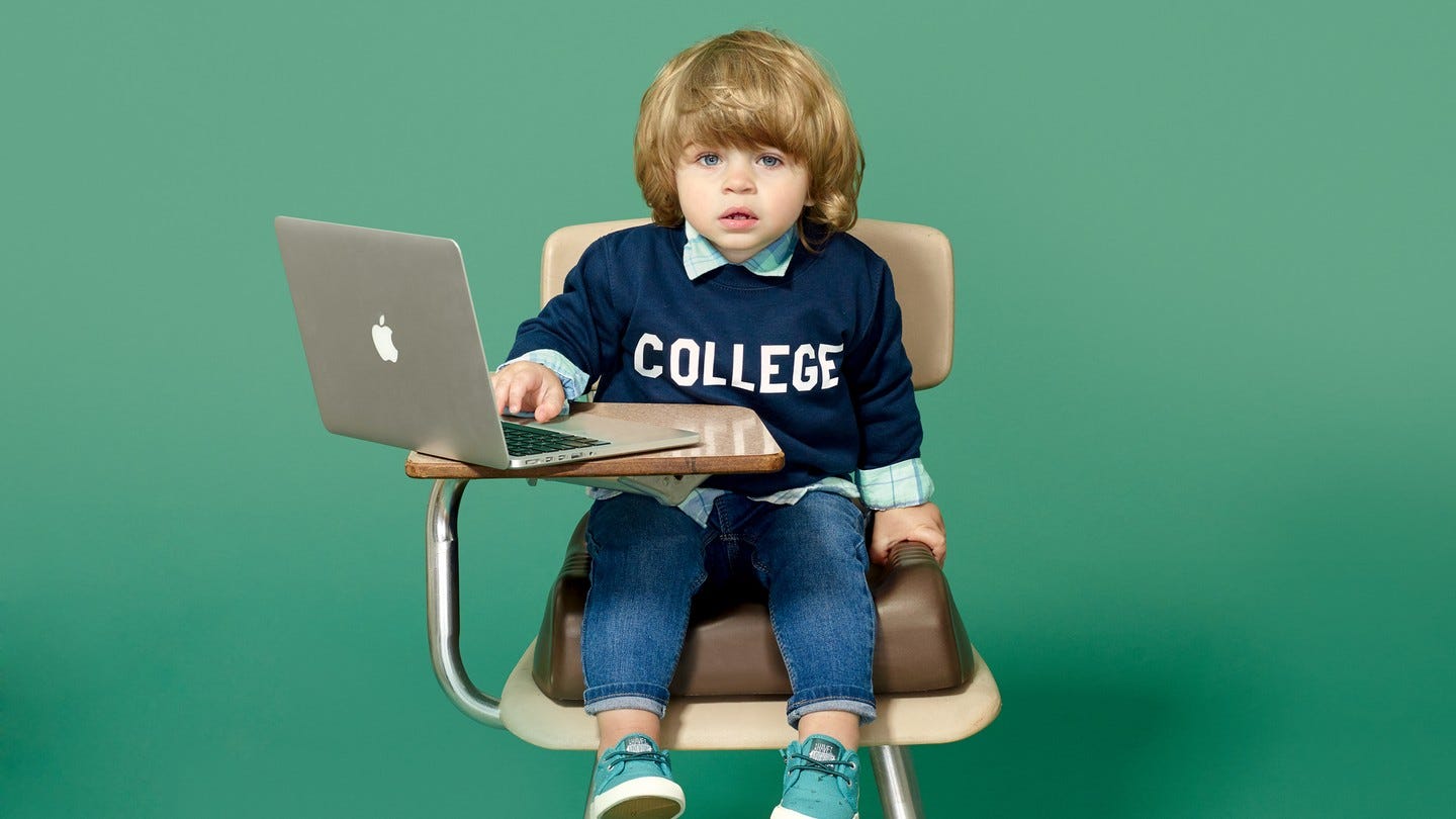 Small child in a desk chair with a laptop, dressed in a collegiate sweatshirt