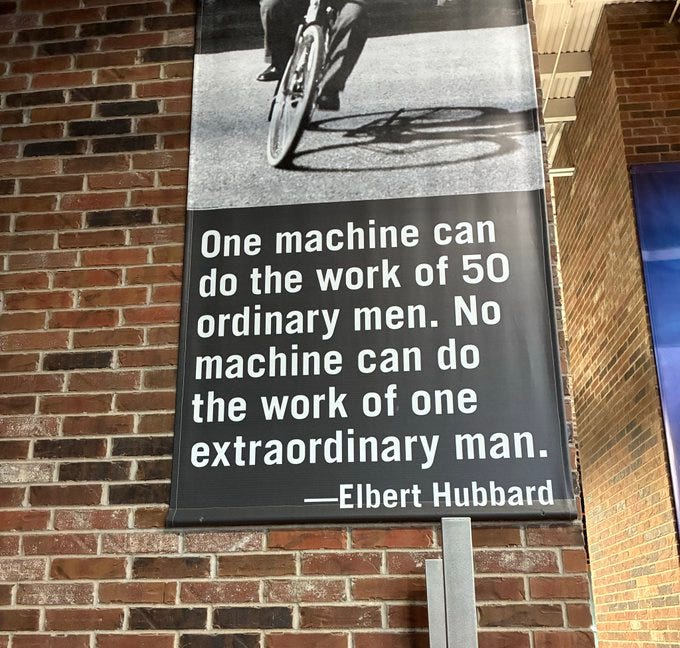 Black and white photograph of a man riding a bicycle on a road, integrated into a poster with bold black text overlay quoting One machine can do the work of fifty ordinary men. No machine can do the work of one extraordinary man. -Elbert Hubbard, set against a brick wall background.