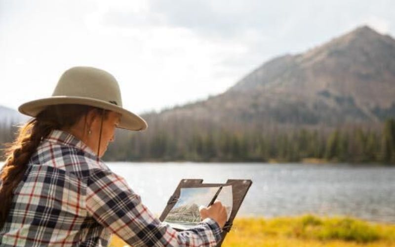 Woman with small easel working on a watercolor painting outside. She is wearing a flannel and wide-brimmed hat. She's over looking golden grass, a mountain lake, and a mountain peak.