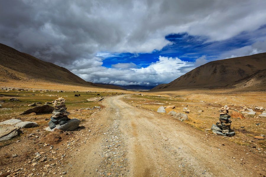 A winding road marked by small stone cairns stretching into the distance beneath a wide unsettled sky. A winding road marked by small stone cairns stretching into the distance beneath a wide unsettled sky.