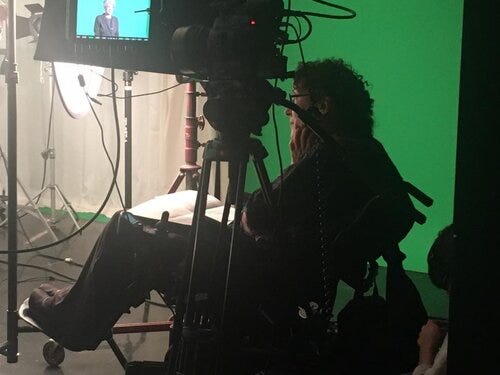 Writer Susan Nussbaum, sitting in a chair by a green screen, as a film is being made in front of her. Writer Susan Nussbaum, sitting in a chair by a green screen, as a film is being made in front of her.
