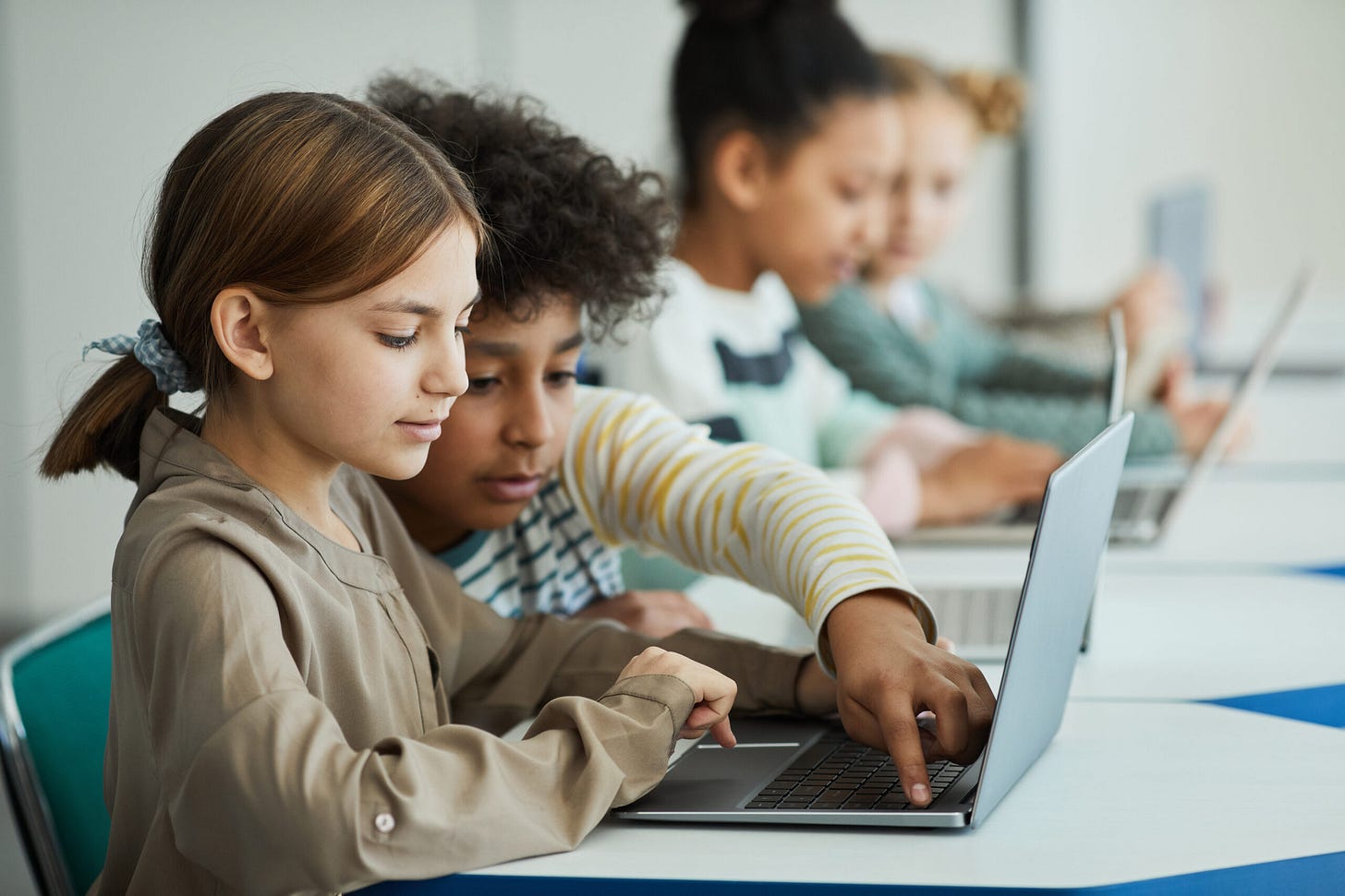 Side view at diverse group of children sitting in row at school classroom and using laptops...