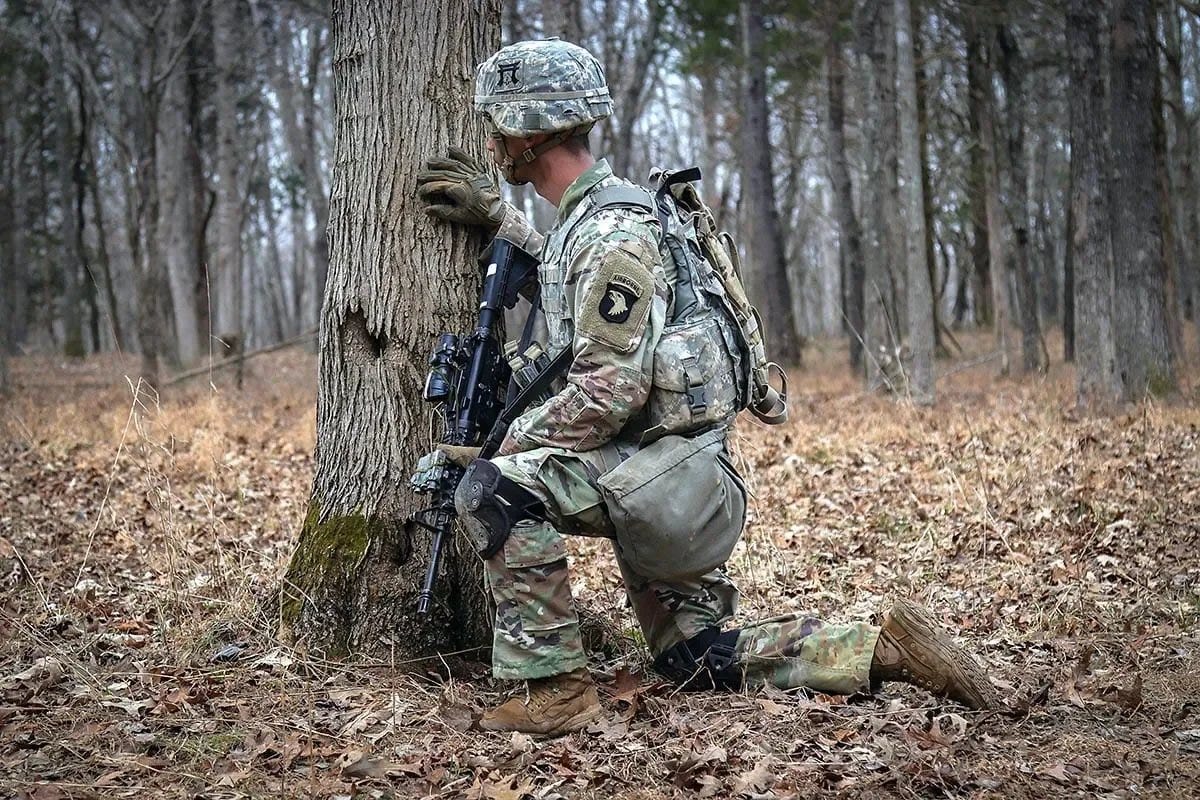 A soldier in camouflage uniform kneeling in a forest, resting against a tree with eyes closed and head bowed.