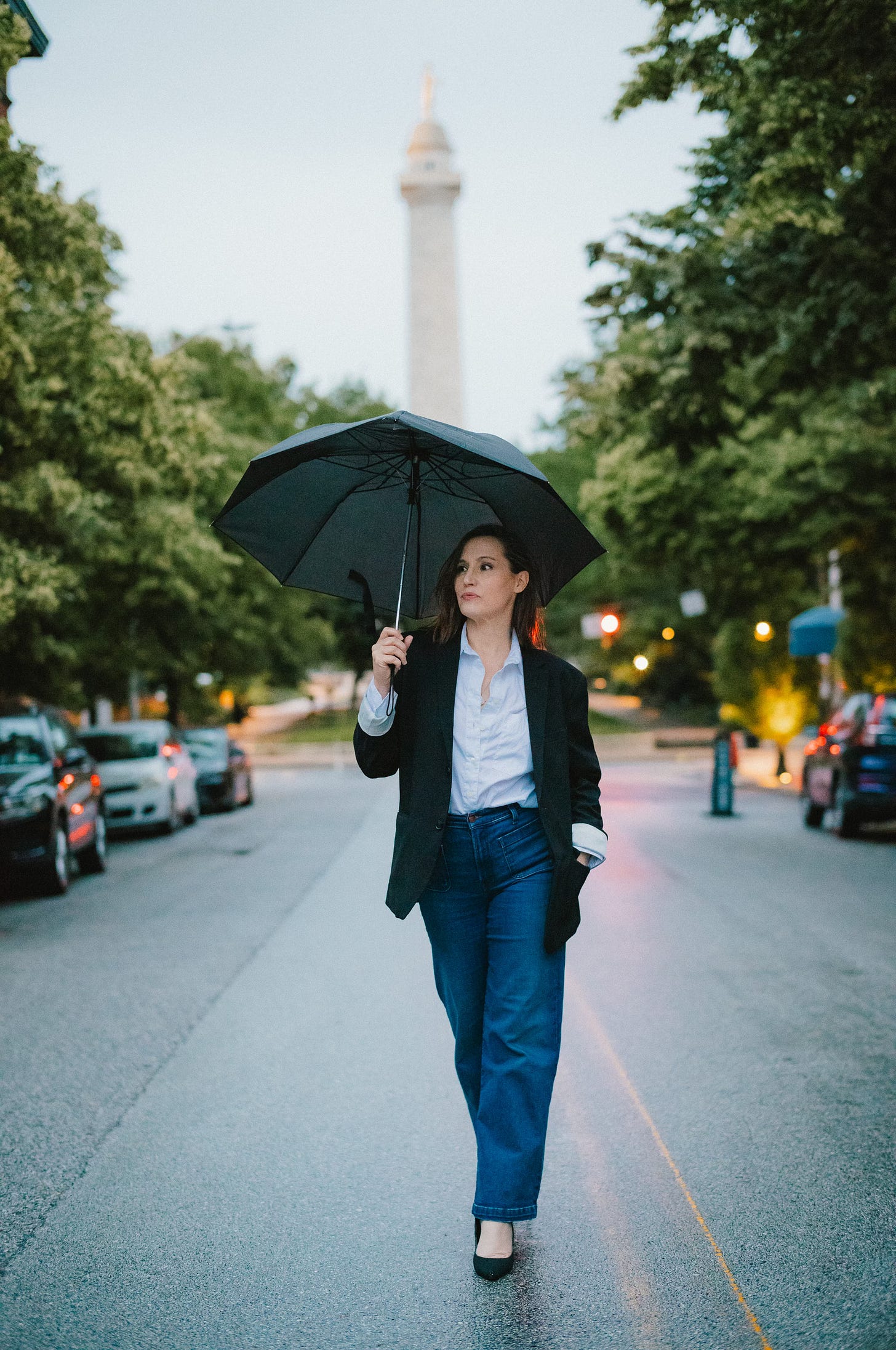 Woman in business casual attire walking confidently down a tree-lined street at dusk, holding a black umbrella, with a monument visible in the background and warm street lights creating a golden glow