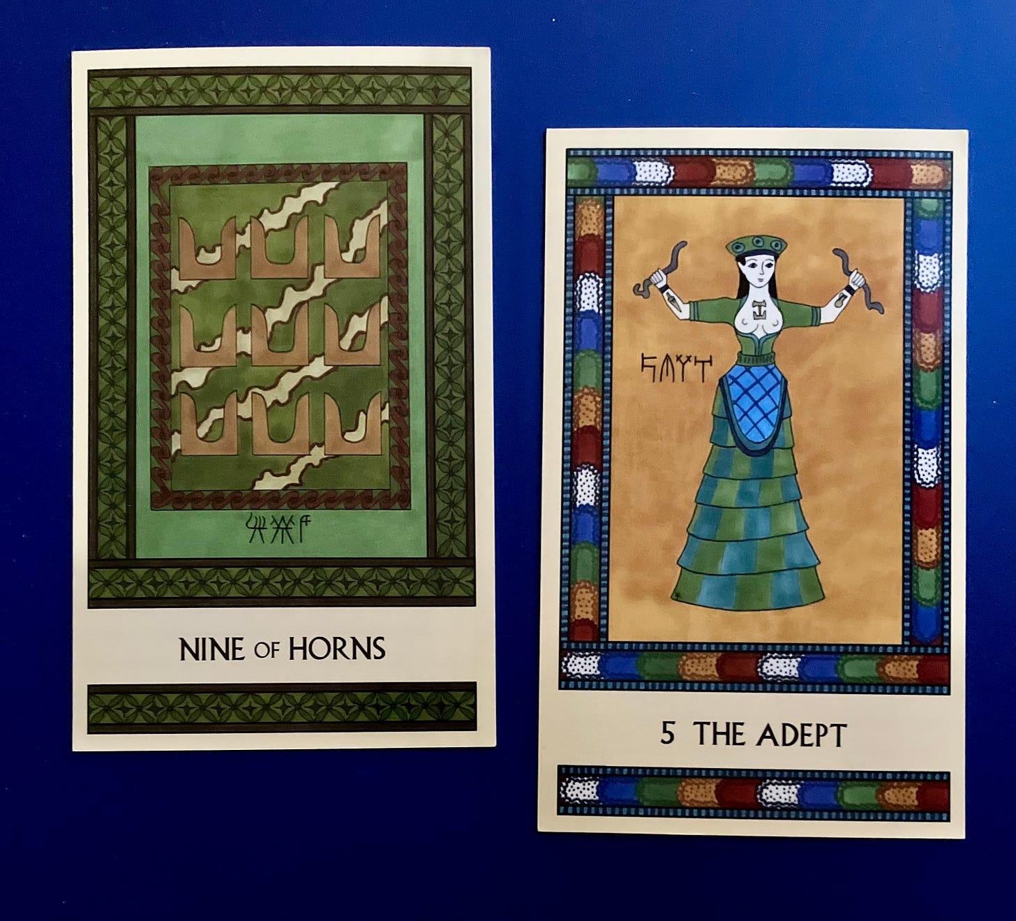  Two Minoan Tarot cards side by side on a dark blue surface. The Nine of Horns is in shades of green and brown. It shows a fancy wall painting depicting nine pairs of sacred horns on a patterned background. The Adept has a multicolored border. It shows the Minoan Snake Goddess figurine, holding a snake in each hand.