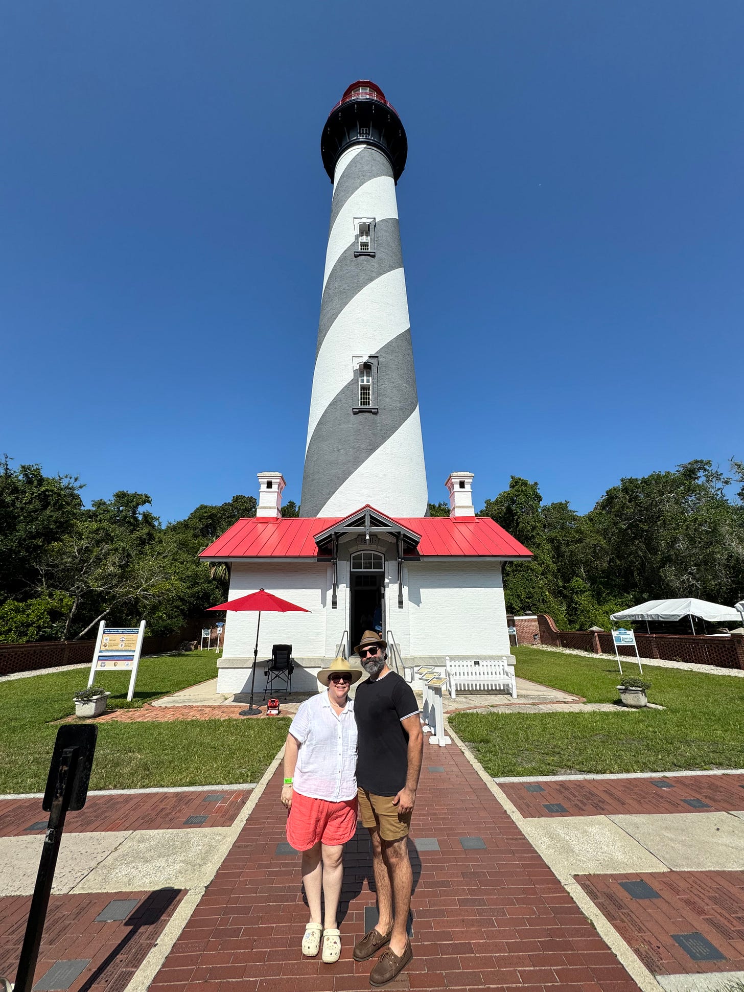 A woman with a white shirt, pink shorts, and beach hat, stands with a bearded man with a blue shirt, khaki shorts, and a fedora, in front of the St. Augustine Lighthouse. A woman with a white shirt, pink shorts, and beach hat, stands with a bearded man with a blue shirt, khaki shorts, and a fedora, in front of the St. Augustine Lighthouse.