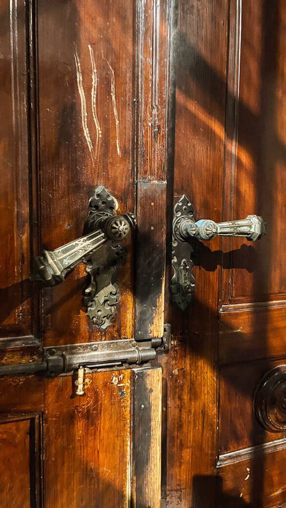 1) Ornate copper handles of a wooden door, turning green from oxidization. One handle is slightly downturned, as if being opened from the other side. 2) Two yellow tulips, mostly closed, poking up through dry dirt. 3) A small pond where a swan swims & a black + grey bird flies overhead through a blue sky. 4) Inside a small restaurant, some small tables & chairs are next to a window that looks out across a small town of hold houses & cobblestone streets. It's night time & there’s a tree of fairy lights next to the tables.