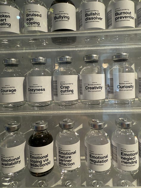 Art installation titled 'Bottled Pondering' by Aepenton, on display at the Design Museum Denmark. The piece features a wall-mounted grid of small glass vials, each labeled with a different abstract or emotional concept, such as 'Inner Critic Softener' or 'Courage Enhancer.' The vials are arranged in neat rows within a transparent display case, evoking the look of a scientific archive or apothecary cabinet of thoughts and feelings.