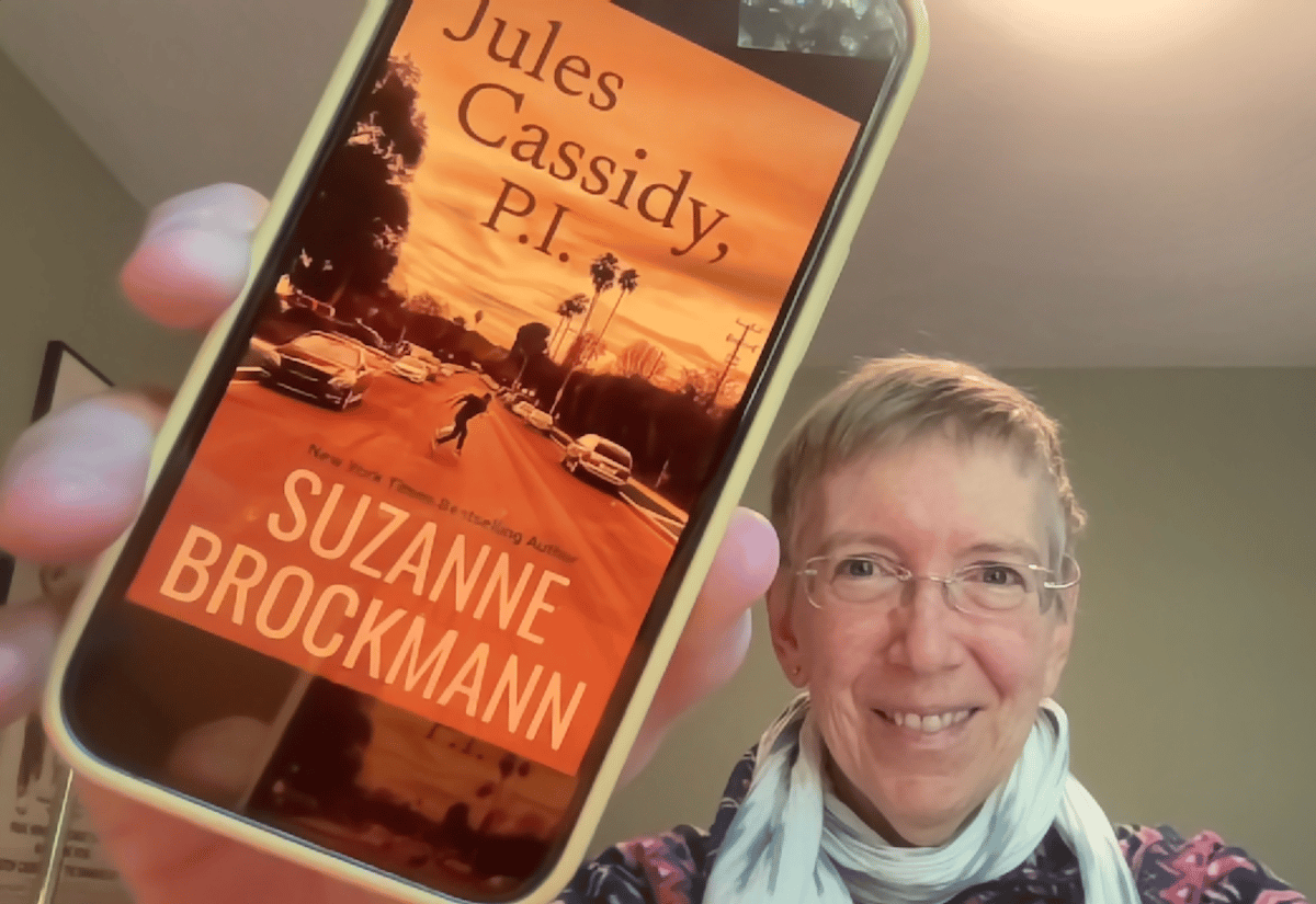 Your author, Suz Brockmann, a tired-looking older white woman with short fair hair and glasses smiles as she holds up her iPhone with the cover art to her latest book, Jules Cassidy, P.I. The cover art shows a reddish-tinged evening sky and a road in a tired-looking Los Angeles Valley neighborhood where the small figures of two men, one large, one smaller, run across the street. Palm trees and electrical poles are silhouette against the sunset.