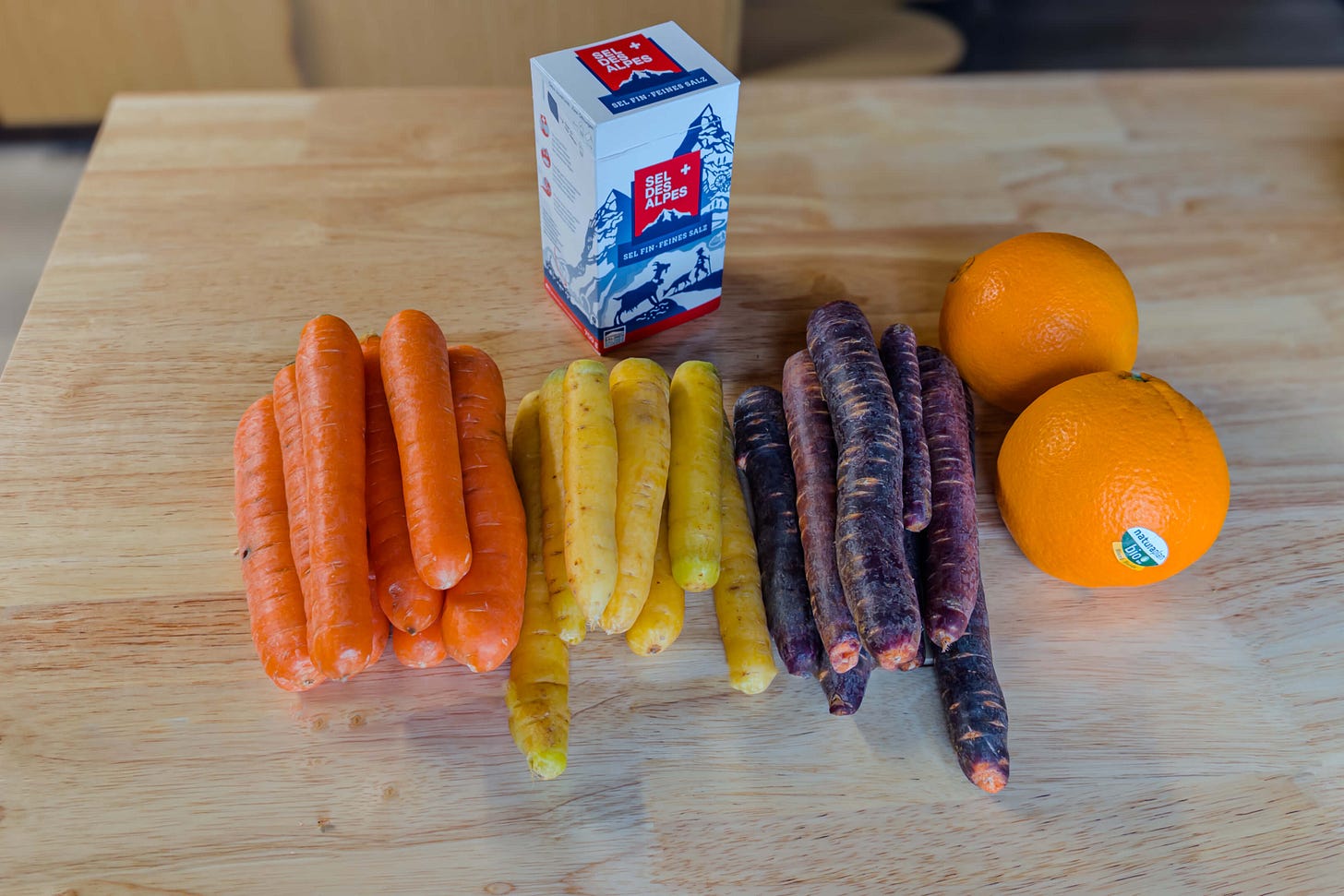 Auf einem Holztisch steht eine Packung «Sel des Alpes», davor liegen verschiedene Karottensorten – orange, gelbe und violette (Purple Haze) – sowie Orangen.