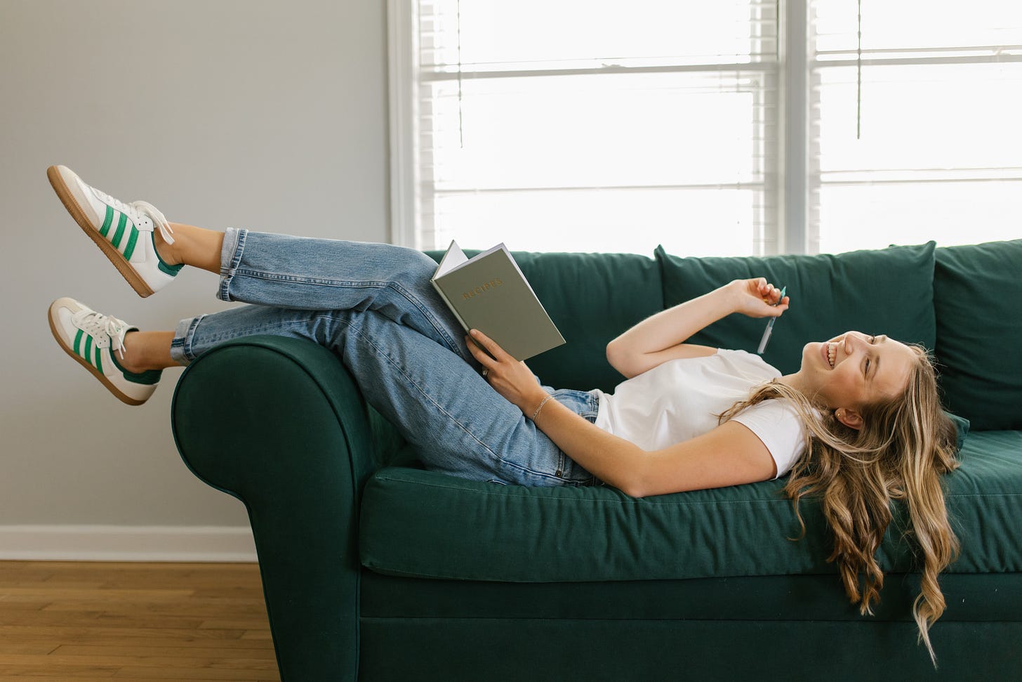 girl in a white shirt with blue jeans and green shoes, leaning back on a green couch writing recipes in her green notebook