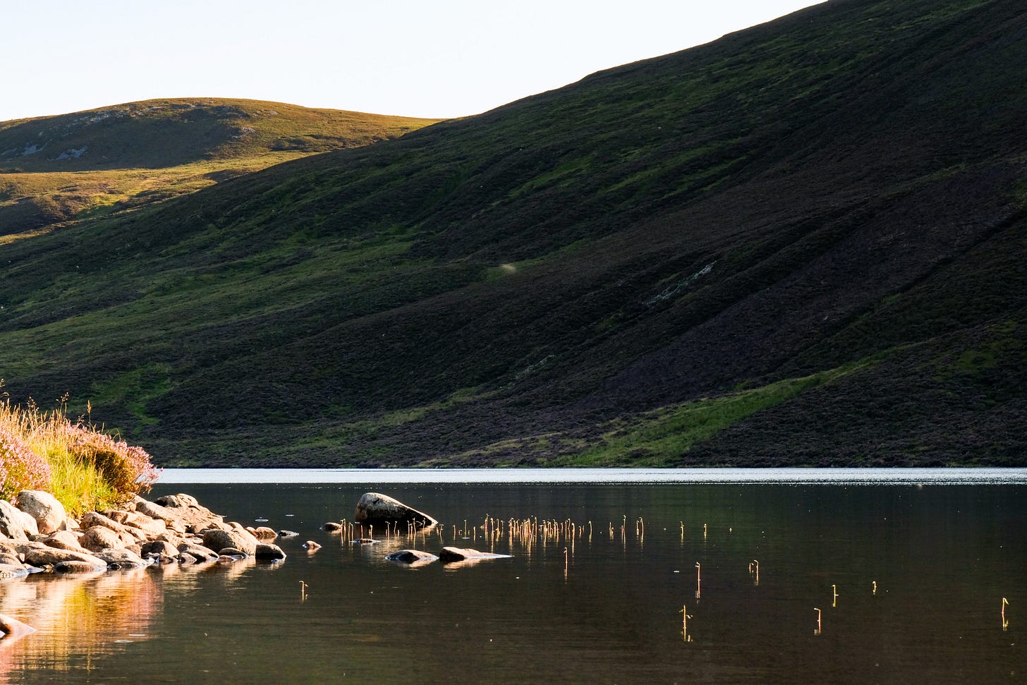 A calm Highland loch at dusk. The sunset touches the water’s western edge, while a steep hillside descends into shadow. Only the top of the hill catches the last golden light.