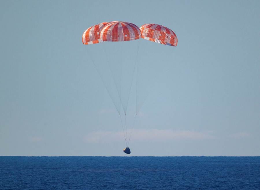 A crew capsule descends under red and white parachutes toward the blue ocean against a blue sky. A crew capsule descends under red and white parachutes toward the blue ocean against a blue sky.