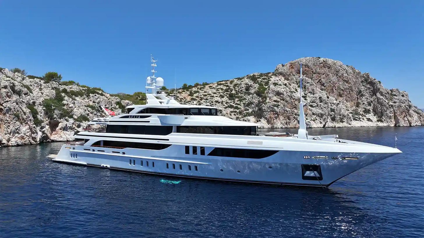 The Aelia, a gleaming Benetti superyacht, anchored near rocky Mediterranean cliffs on a bright summer day.