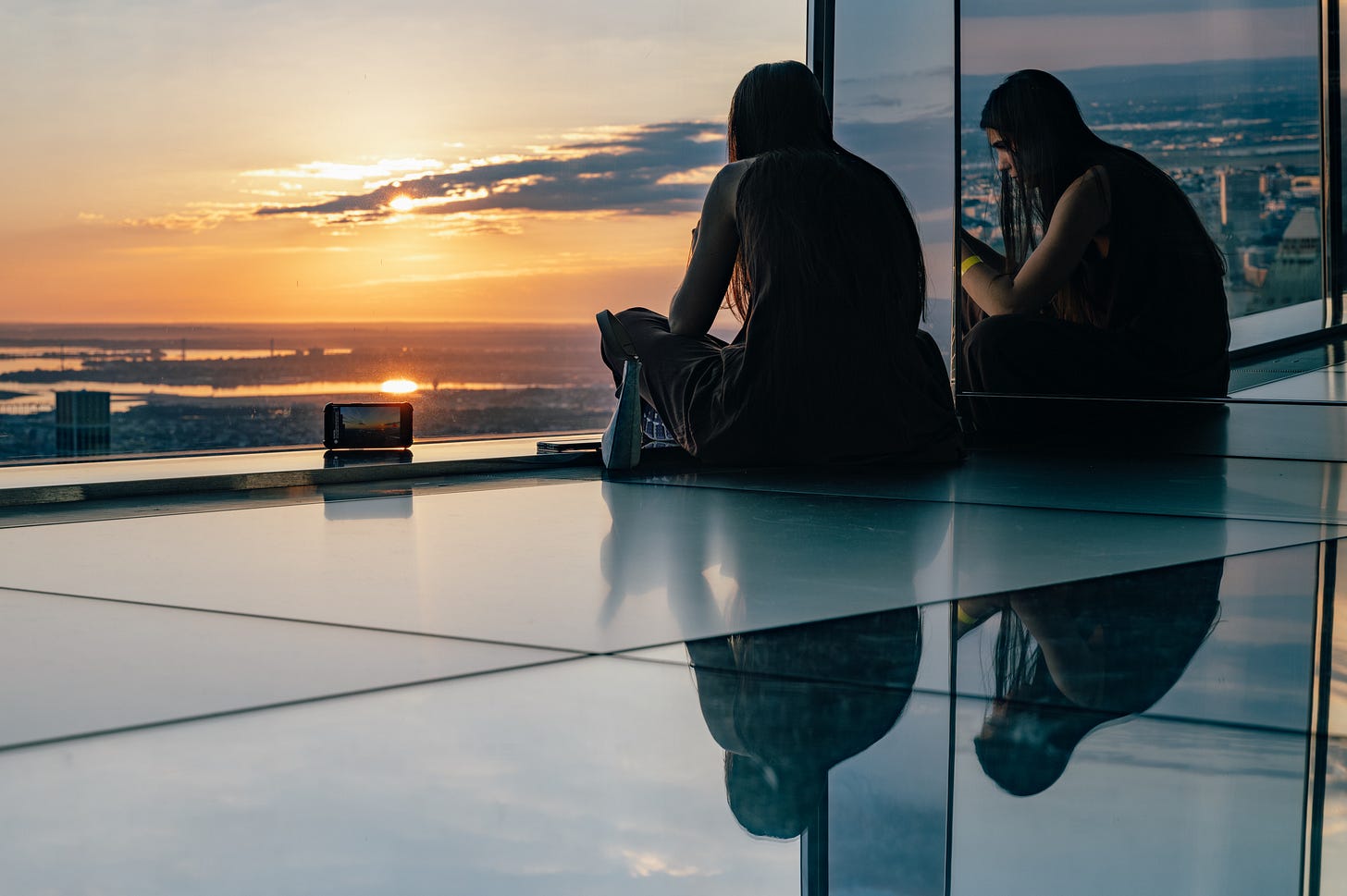 A woman sitting on a mirrored floor reflected in a mirrored wall watching the sun rise over the water, her reflections beneath her and a phone propped up on the floor. A woman sitting on a mirrored floor reflected in a mirrored wall watching the sun rise over the water, her reflections beneath her and a phone propped up on the floor.