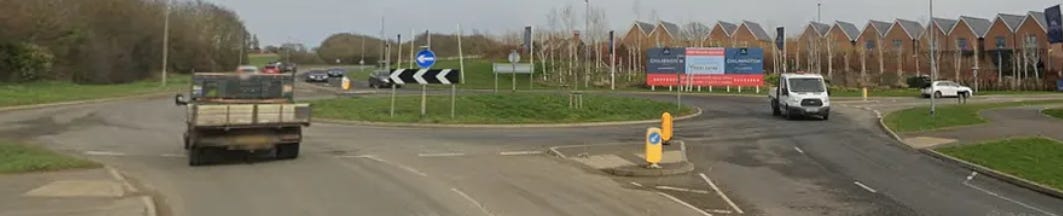 A roundabout with pedestrian tiles.  2 lanes then a small refuge area and another 2 lanes of traffic.  Very close to the roundabout. Several roads converge, bordered by grassy verges and modern brick houses under an overcast sky.  Vehicles including a truck and a white van travel around the roundabout whilst roadside banners advertise new housing developments.