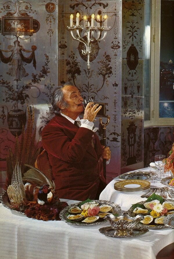 Salvador Dalí seated at an opulent dining table, raising a glass in a velvet jacket, surrounded by elaborate silver platters of food, ornate wallpaper, and candlelight—evoking the theatrical surrealism of his cookbook, Les Dîners de Gala.