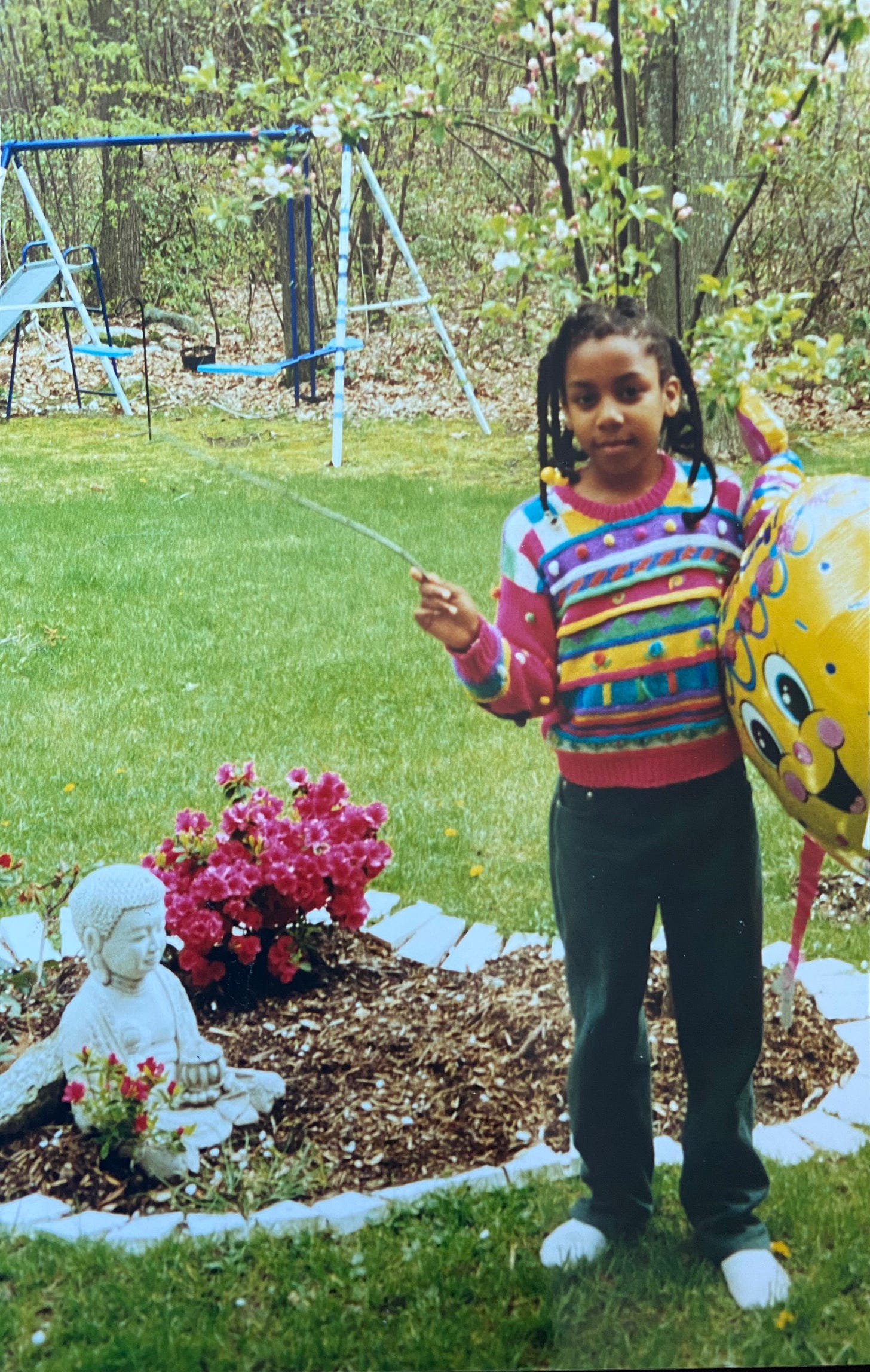 Brown-skinned girl child standing in a springtime backyard garden next to a small rose garden and a buddha statue. She is wearing braids with barettes, a pink colorful sweater, green jeans, and she is holding a large yellow birthday balloon.   