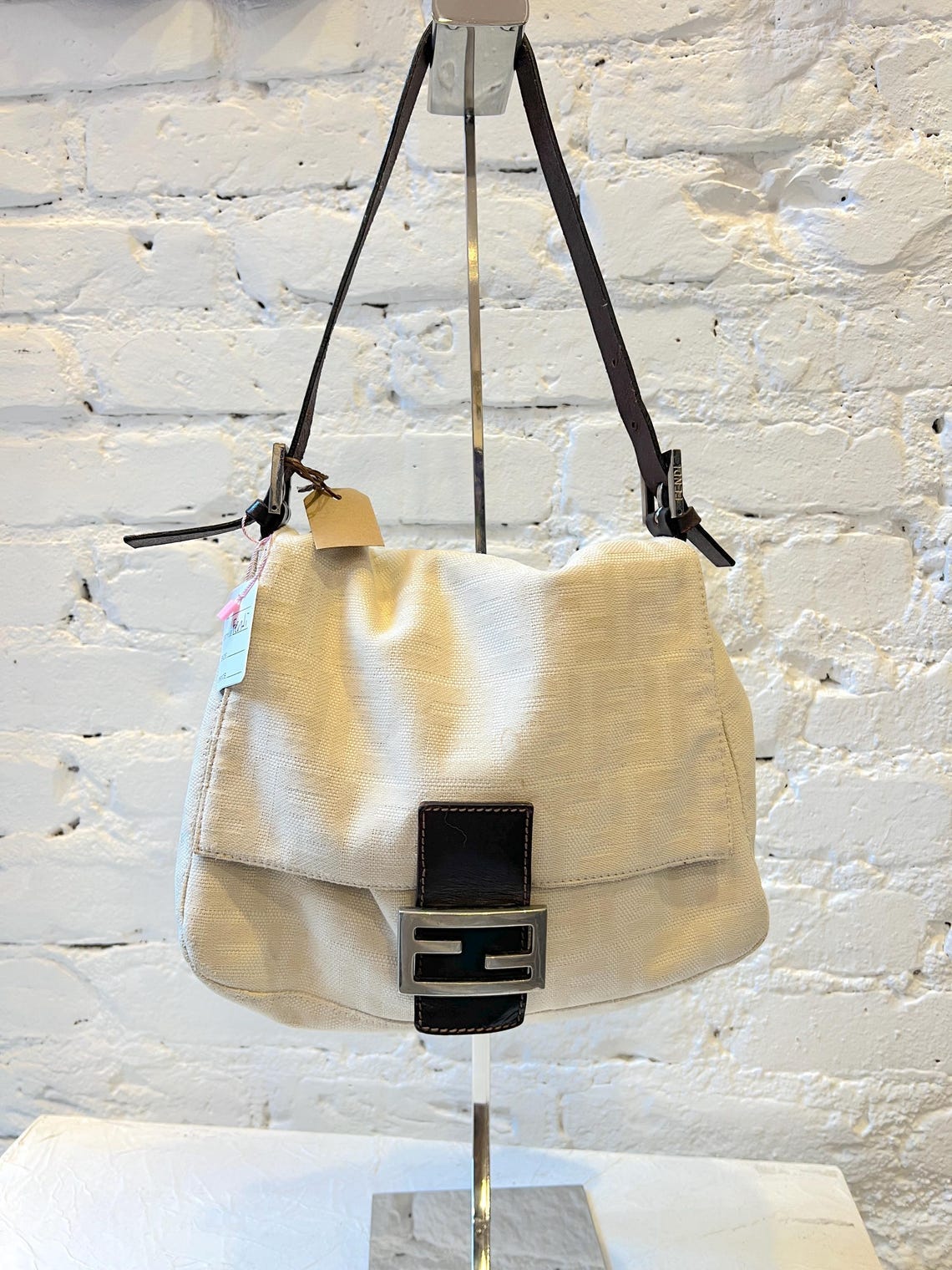 May include: A white canvas shoulder bag with a brown leather strap and a silver Fendi logo. The bag is hanging on a silver stand against a white brick wall.