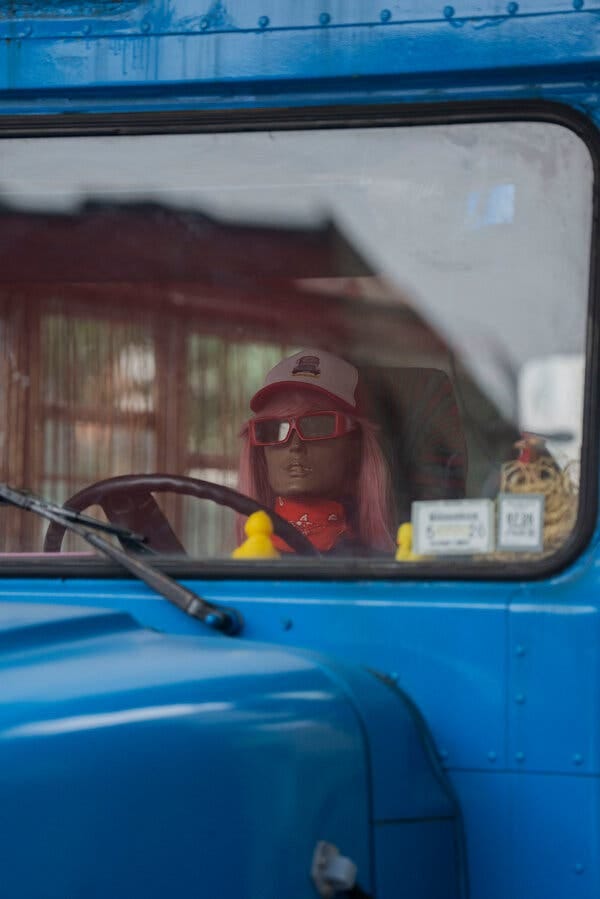 A mannequin wearing a colorful wig, sunglasses and scarf sits behind the wheel of a blue bus.