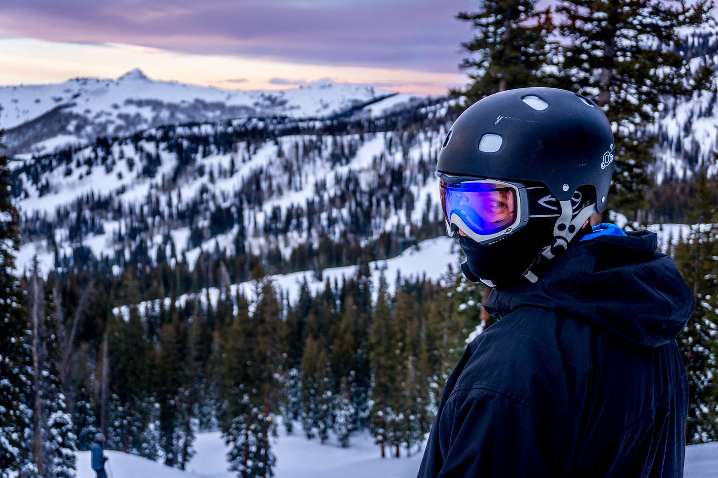 Skier in helmet and goggles looking over a snowy mountain valley at dusk Skier in helmet and goggles looking over a snowy mountain valley at dusk