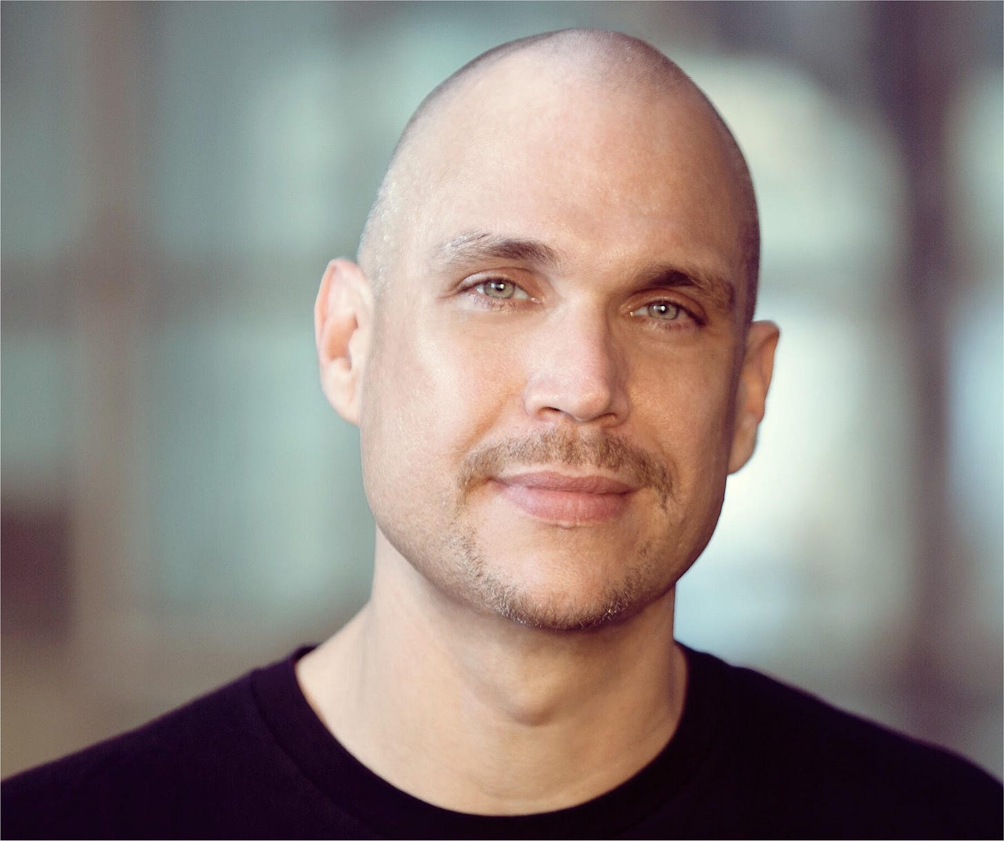 Man with a goatee and light-colored eyes looks a the camera with a slight, closed -lip smile. He wears a black t-shirt.