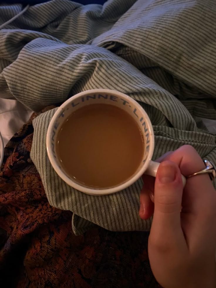 This may contain: a person holding a cup of coffee on top of a bed next to a blanket