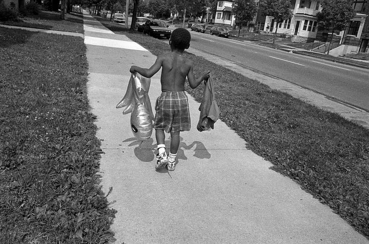 Two photographs. One is of two boys looking at large format photos posted in a store window on Lake Street. The second is a photo from the exhibit. It shows a small boy walking down the street holding a balloon and his tee shirt.