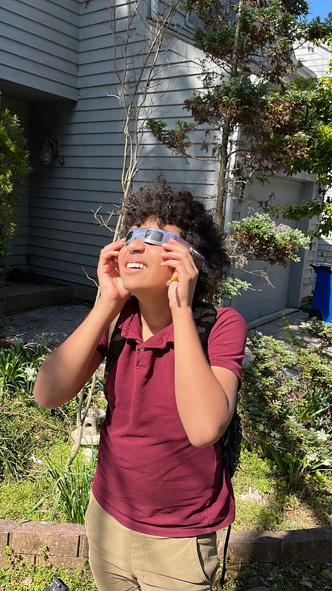 A gray tabby cat sits with front paws on the floor. A boy child with dark curly hair squints at the solar eclipse through eclipse goggles, holding his hands to the edges of the goggles. A sunset is visible through spring flowering trees. A pile of framed posters and suitcases and other belongings sits in the center of a room. Another gray tabby with white paws holds the hand of a human and looks into the camera. A lake's surface glitters in the sun behind a black wrought iron fence. A magnet and a post-it note with the words May 9th are stuck to a stainless steel surface. A human hand holds a crochet hook above a purple crocheted scarf. A pin reads, Matzah Balls Not Border Walls.