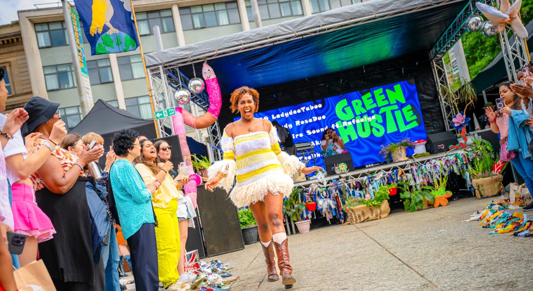 A crowd of people watching a woman in a white and yellow dress walk down the catwalk, with a stage with a sign that says 'Green Hustle' behind her A crowd of people watching a woman in a white and yellow dress walk down the catwalk, with a stage with a sign that says 'Green Hustle' behind her
