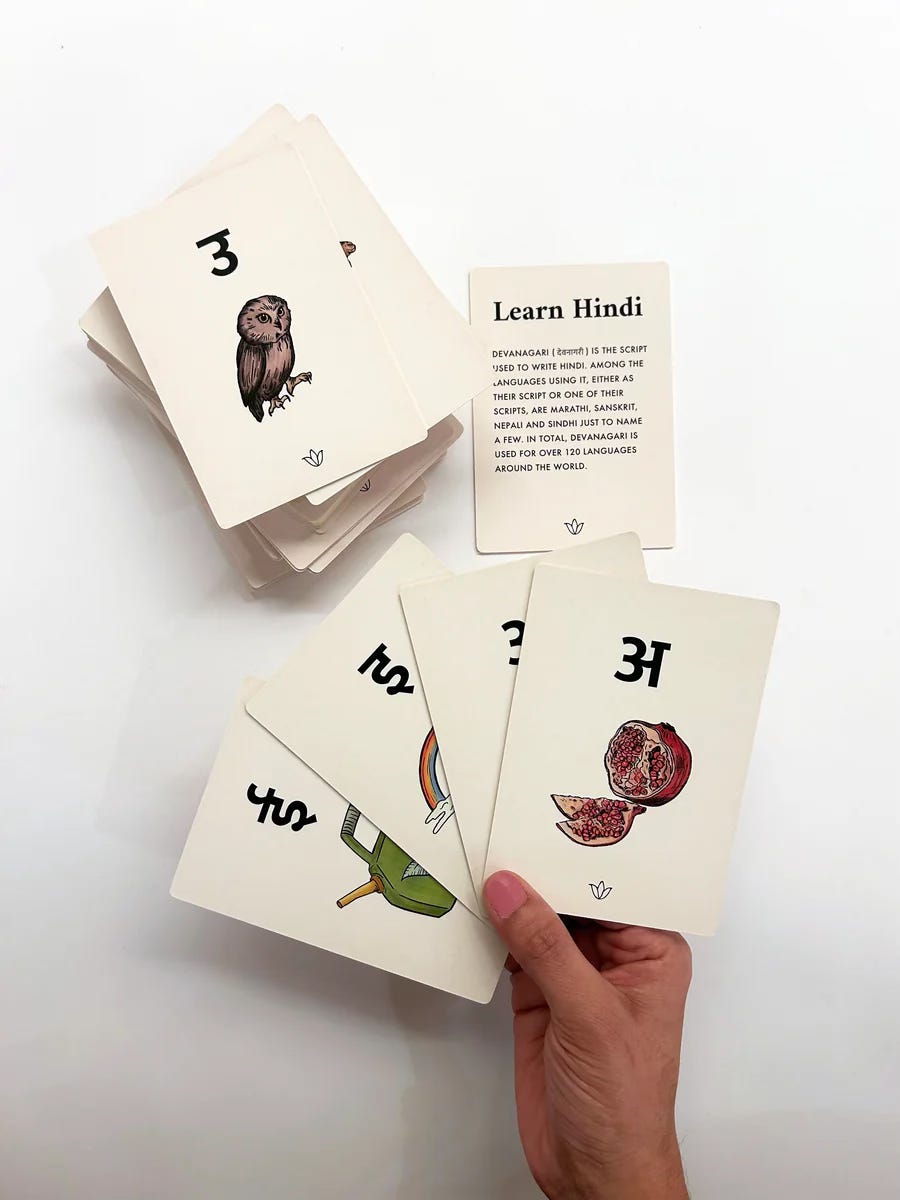 Here's alt text for this image: "A hand holds three flashcards from a Hindi learning deck. The visible cards show Devanagari script with illustrations: a chili pepper, a pomegranate cut in half, and a question mark card. Behind them is a stack of additional cards, with the top card displaying a detailed illustration of an owl labeled with the number 3. An informational card titled 'Learn Hindi' explains that Devanagari script is used to write Hindi and over 120 other languages worldwide