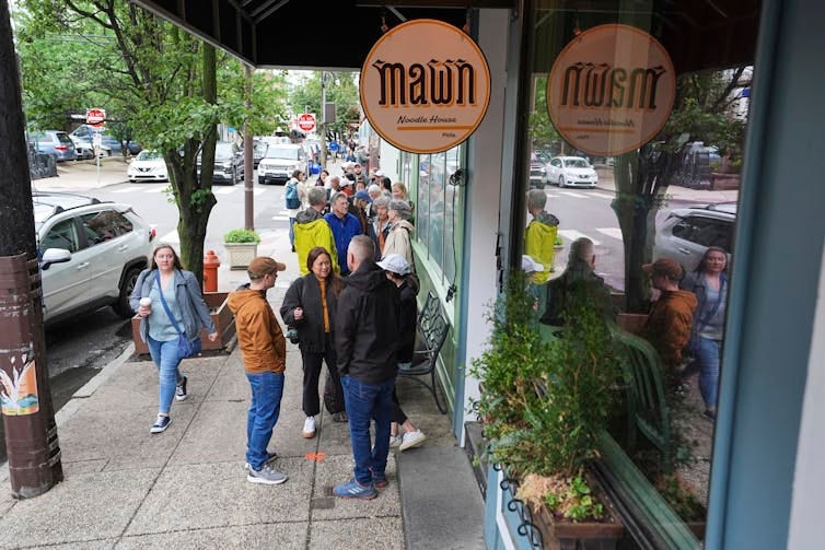Line of people form on sidewalk outside a business with a circular sign that says 'Mawn'