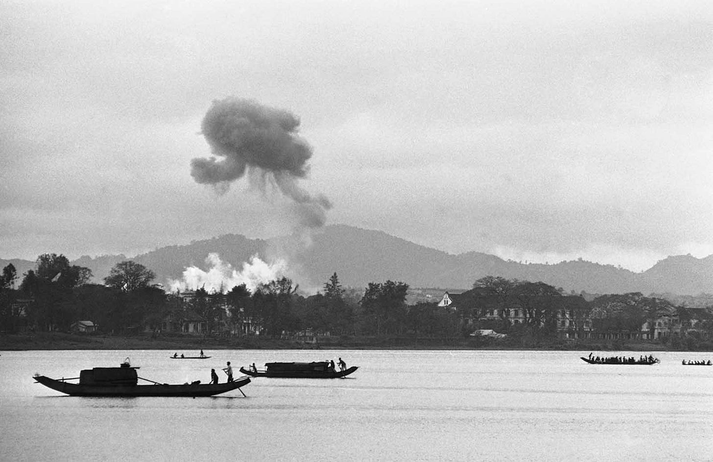 Napalm air strikes ignite the skies above a village near Hue on February 28, 1963, as houseboats navigate the Perfume River, illustrating the intense early conflict around the Imperial City.