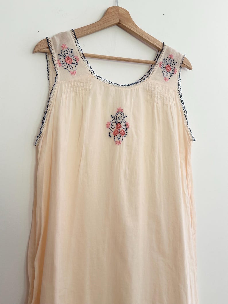 May include: A sleeveless cream-colored nightgown featuring a scoop neckline and dark blue scalloped trim. The garment is decorated with delicate floral embroidery in shades of pink and dark blue on the chest and shoulders, hanging on a wooden hanger.