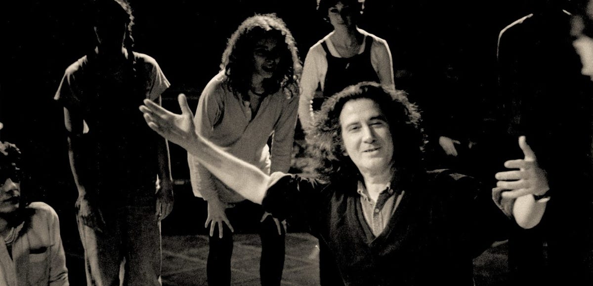 Black-and-white photograph of Brazilian theater practitioner Augusto Boal smiling with his arms extended toward a circle of young workshop participants gathered in a dim rehearsal space in Paris in 1983.