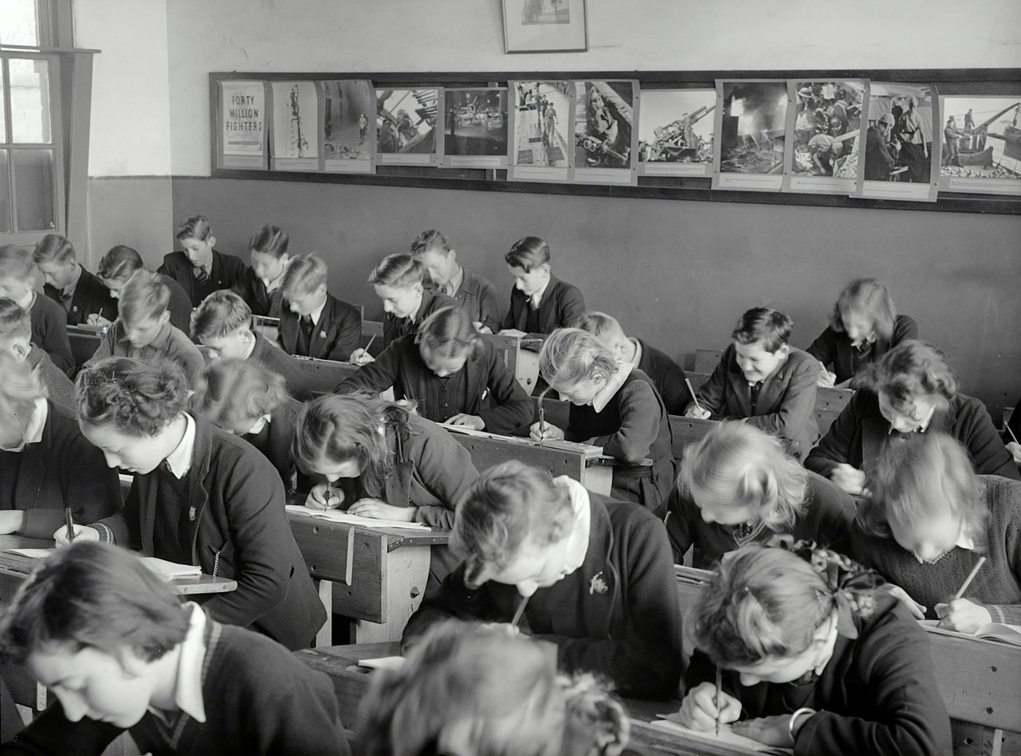Historical black and white photo, perhaps from the 1930s or 40s of a classroom. They are many young people bent over their desks writing in books.