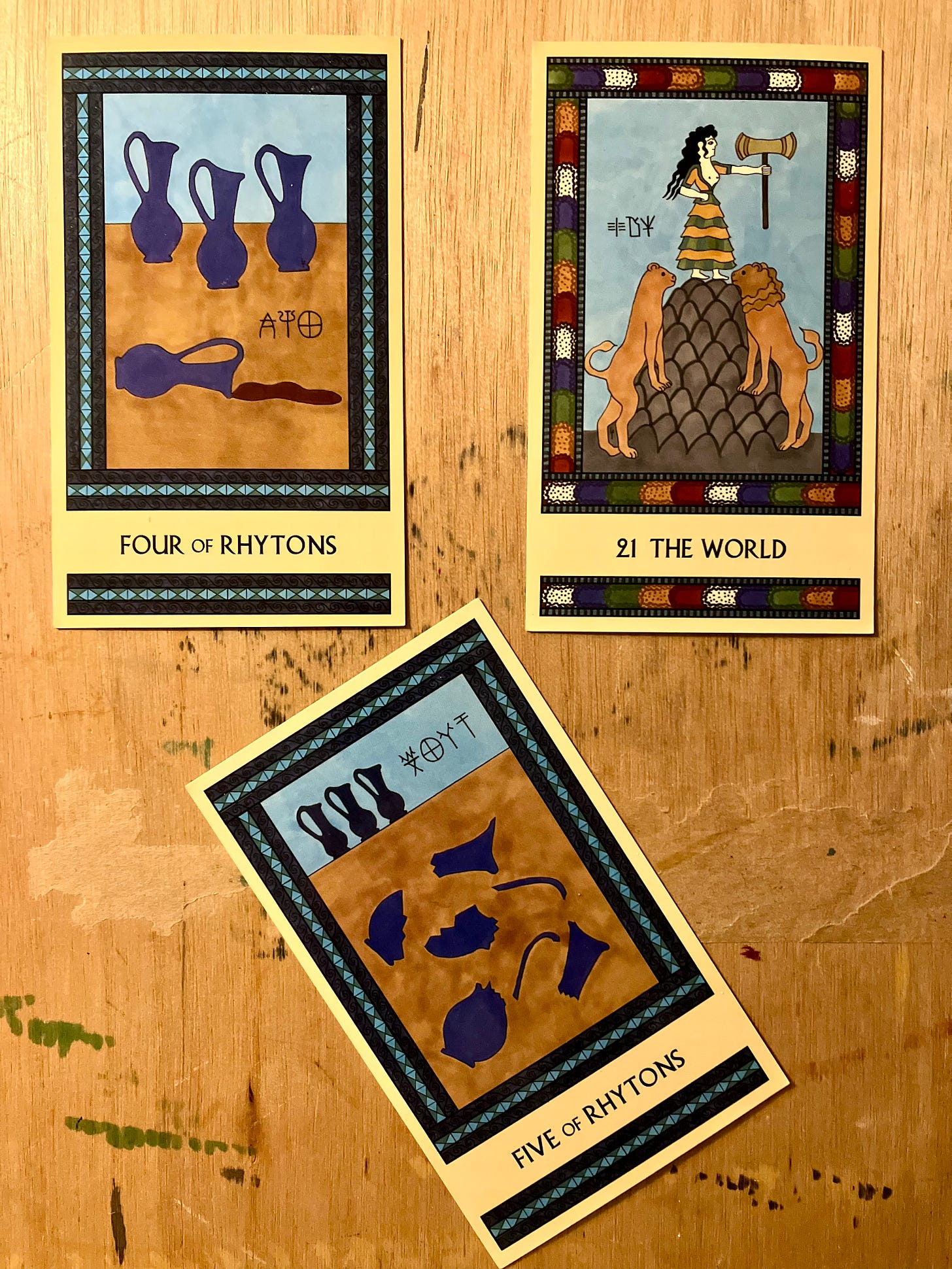 Three Minoan Tarot cards, two side by side and a third at an angle below them, all on a paint-stained wood surface (my art desk). The Four of Rhytons is painted in shades of blue. It shows three blue pitchers standing on a tabletop, with a fourth pitcher tipped over on its side with wine spilling out of it. The World has a bright multicolored border. It shows a Minoan goddess standing atop a tall mountain, holding a labrys. A pair of lions stand on either side of the base of the mountain. The Five of Rhytons is painted in shades of blue. It shows two blue pitchers lying shattered in pieces in the foreground while three more pitchers, still intact, stand in a row in the background. 