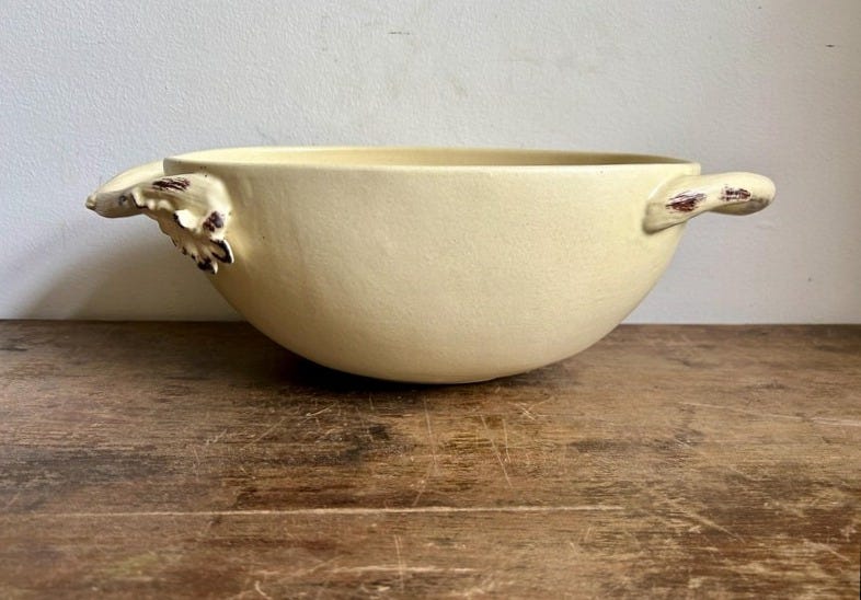 May include: A cream-colored ceramic bowl with two decorative handles. The handles have a leaf-like design with brown accents. The bowl is set on a wooden surface, and the background is a plain white wall. This bowl is suitable for serving food.